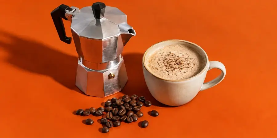 Una moka, una tazza di bevanda ai funghi e una manciata di chicchi di caffè su fondo arancio bruciato