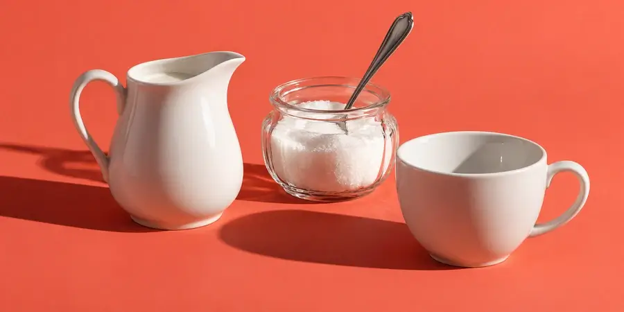 Bricco di latte, zuccheriera di vetro con un cucchiaino e tazza vuota su fondo corallo